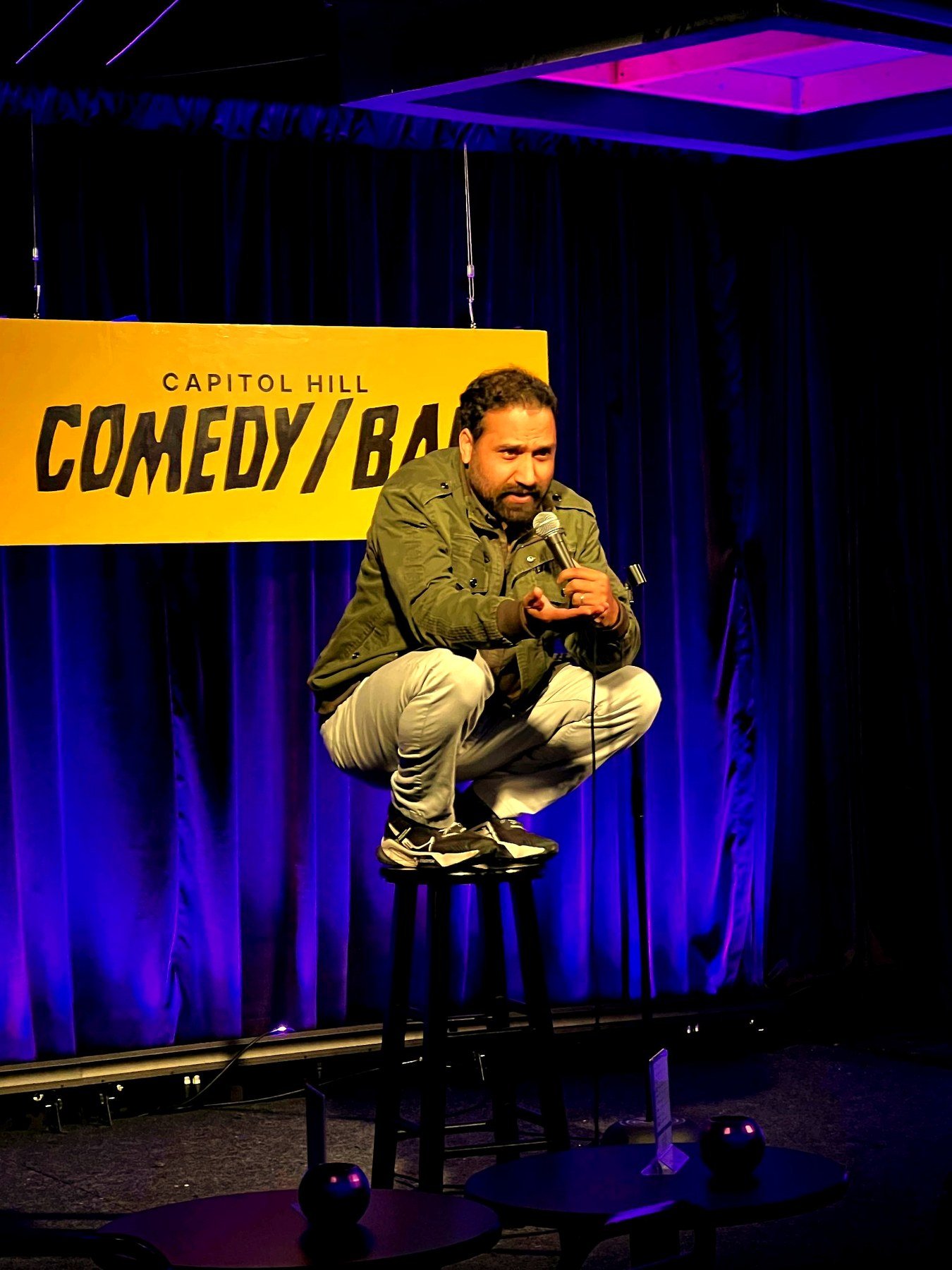 Ish performing live on stage, crouched on a stool with a microphone at Capitol Hill Comedy Bar in Seattle
